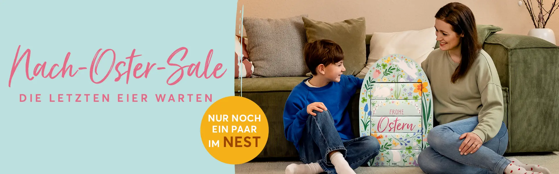 Mutter und Sohn sitzen in einem gemütlichen Wohnzimmer vor dem Sofa auf dem Boden. Zwischen ihnen steht das bunt dekorierte, riesiges brandnooz Osterei mit der Aufschrift "Frohe Ostern". Das Ei ist fast genauso groß wie der etwa 8 jährige Junge.